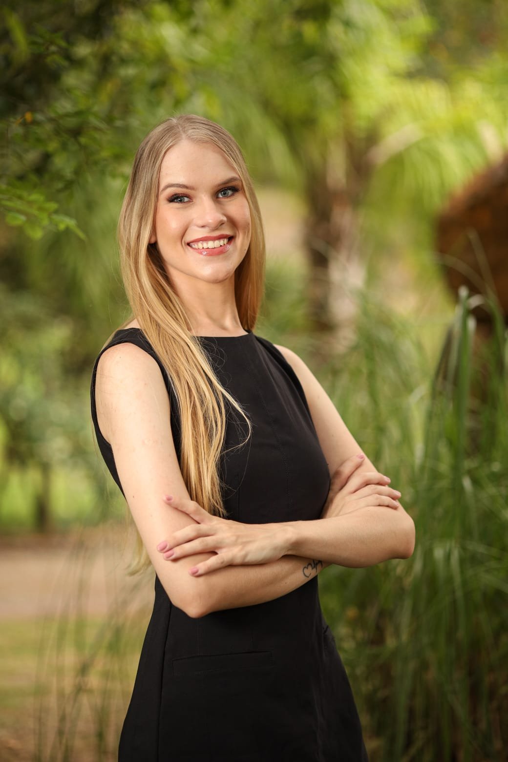 Maíra Becker de braços cruzados, sorrindo, em retrato ao ar livre com fundo de folhagem.