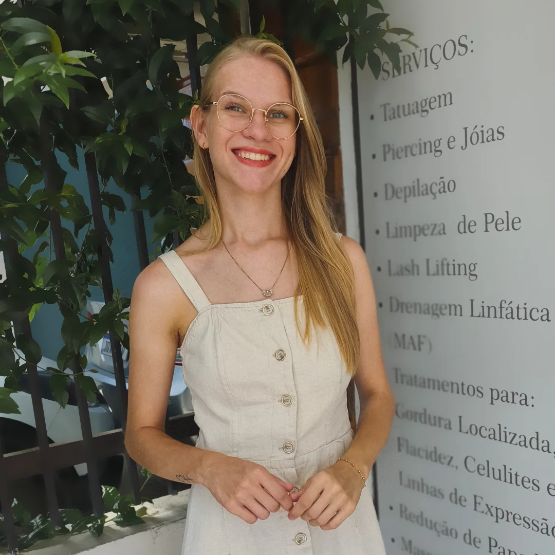 Maíra Becker sorrindo na entrada do Studio Art e Estética, ao lado do painel com a lista de serviços.